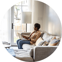 A woman sitting on a couch in her living room with a laptop on her lap, she is looking out the window