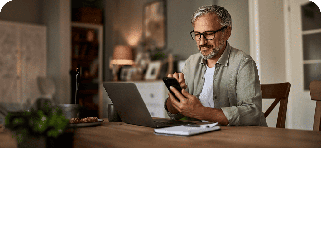 Older man sitting at a table with a laptop while he looks at his phone